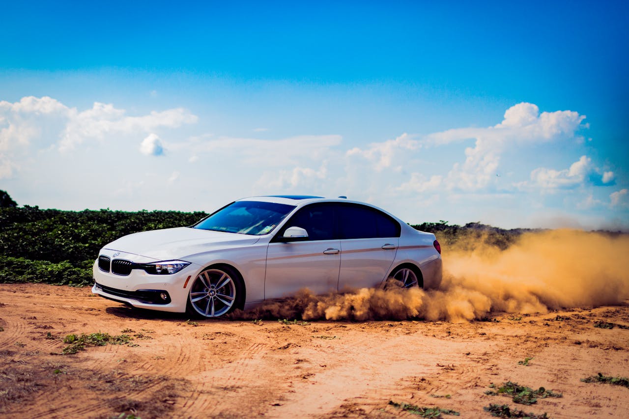 our-services-1 White luxury car drifting on a dirt road, creating a dramatic dust cloud under a blue sky.